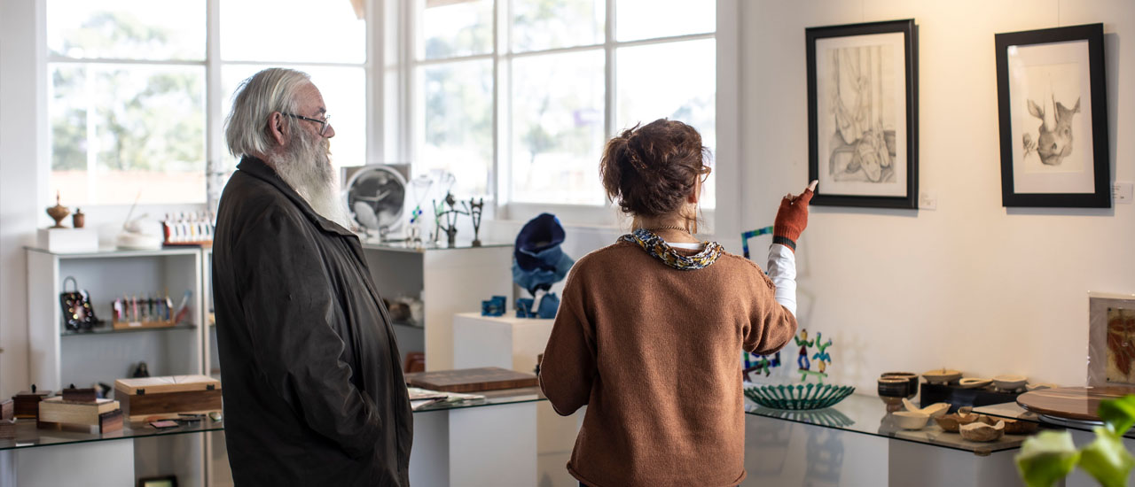 Two people are looking at paintings on a wall inside a small art gallery. Sculptures and other artworks are on display in the background. 
