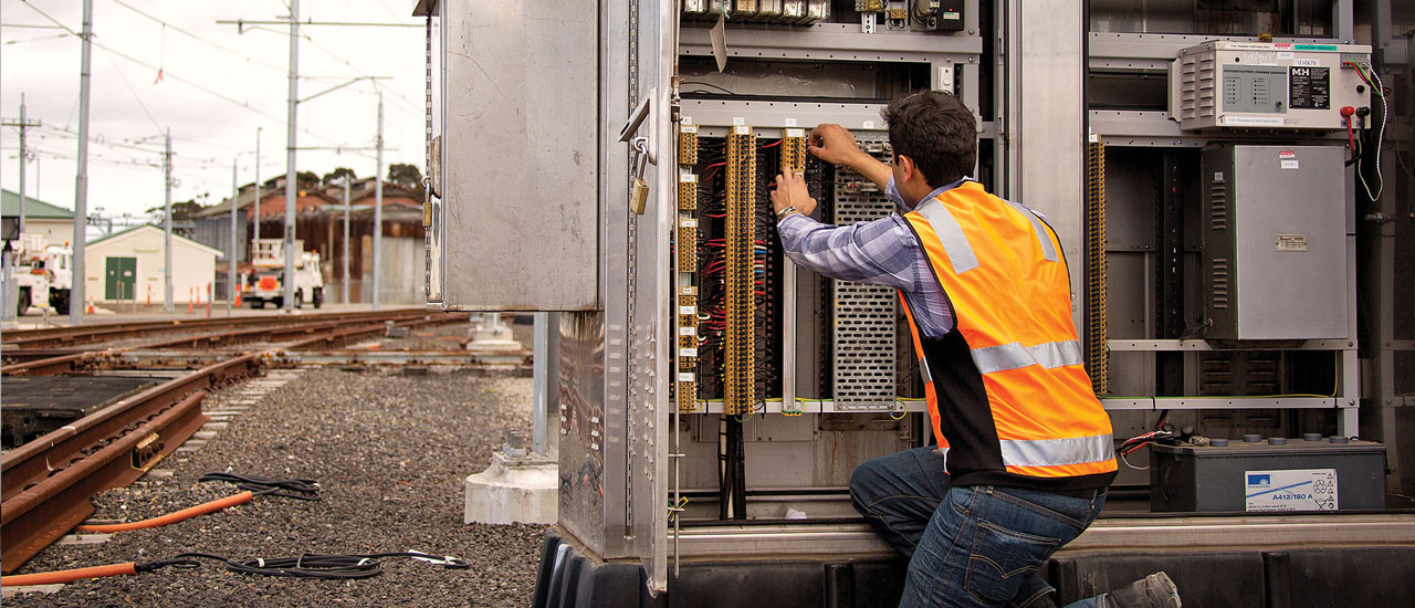 An engineer wearing an orange high visibility vest is installing telecommunications cable in a cabinet beside railway tracks