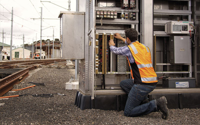 An engineer wearing an orange high visibility vest is installing telecommunications cable into a cabinet beside railway tracks
