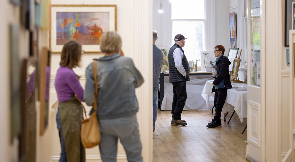 Two rooms in a community arts gallery. Two people in the foreground are looking at an artwork on a white wall. In the background in the second room, two people are surrounded by art, and they are talking to each other.