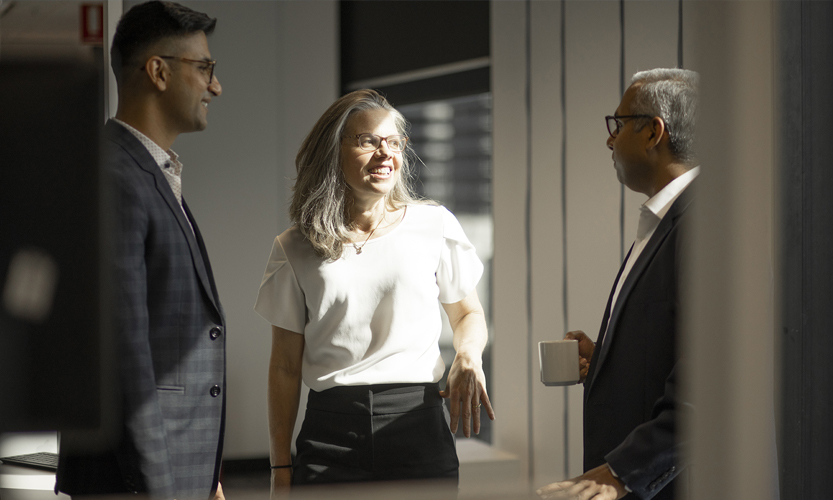 Three office workers wearing suits standing near a window and talking.