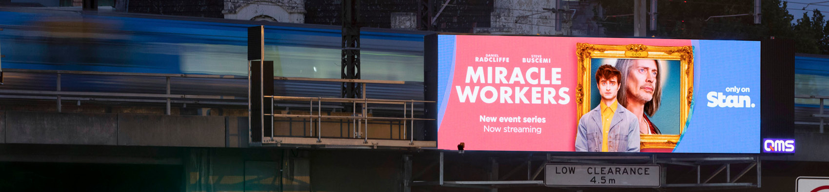 A billboard showing an ad for a TV show streaming on Stan. In the background is a rail bridge with a train passing through it.