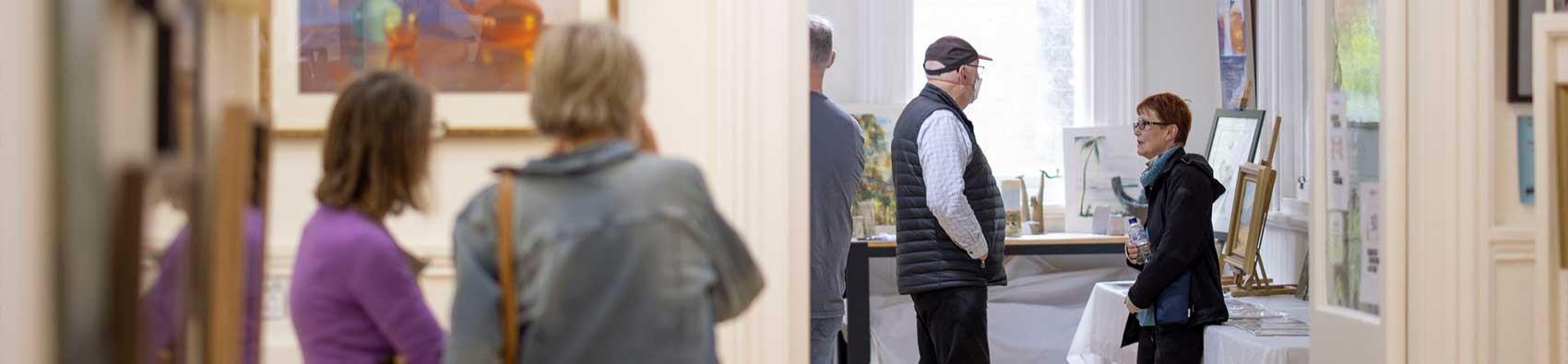 Four people standing in a gallery. Two people in the foreground viewing a painting and two people in the background speaking with each other.