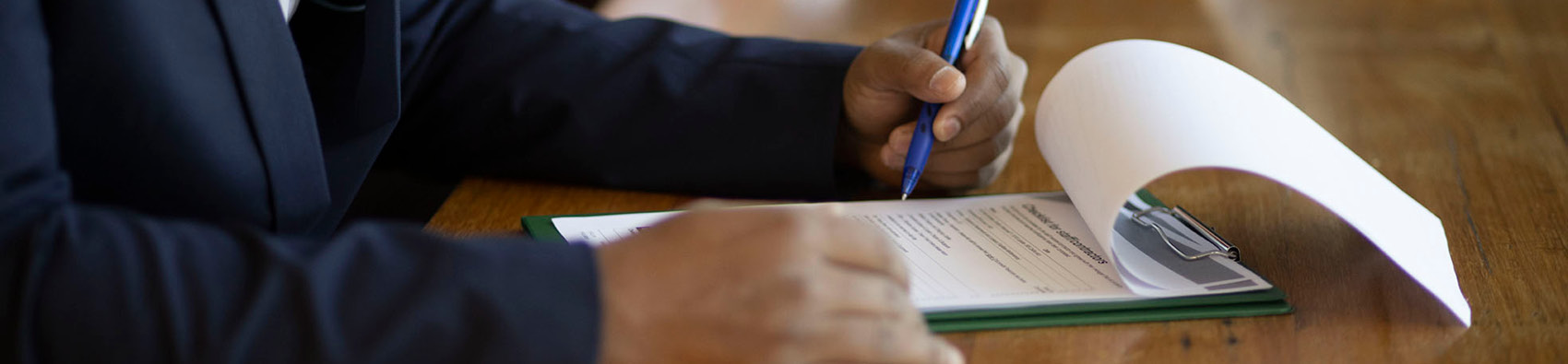 Hands on a desk. One hand is holding a pen against a document.