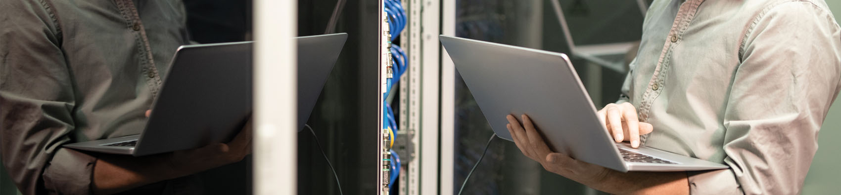 A cropped view of a worker in a server room. The worker is holding an open laptop.