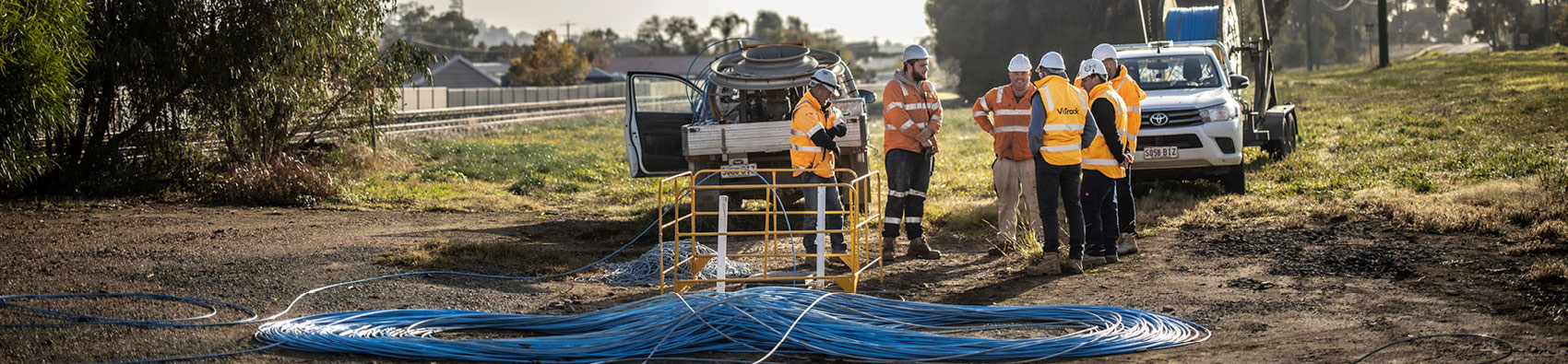 A group of men wearing hi vis safety gear and white helmets. They are standing in field. In the foreground is a big circle of blue cables. In the backrgound there are two white vehicles.