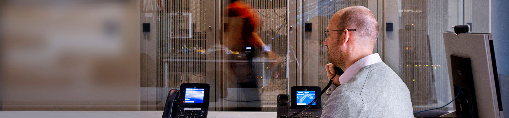 A person sitting at a desk with two phones. The person is talking on one phone. In the background, there are telecommunication servers with someone walking past.