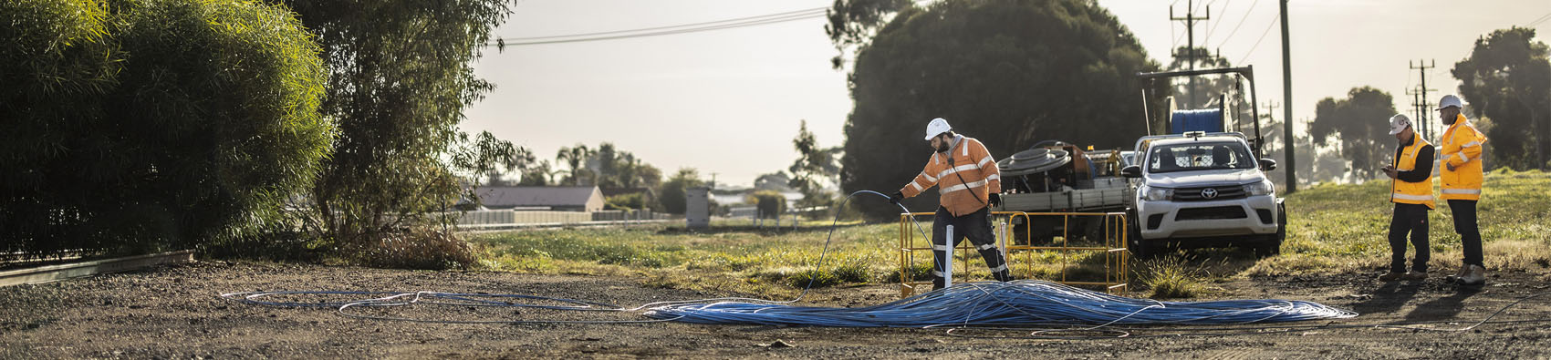 Three wokers in hi vis jackets and hard hats working in a field with a truck in the backrgound.