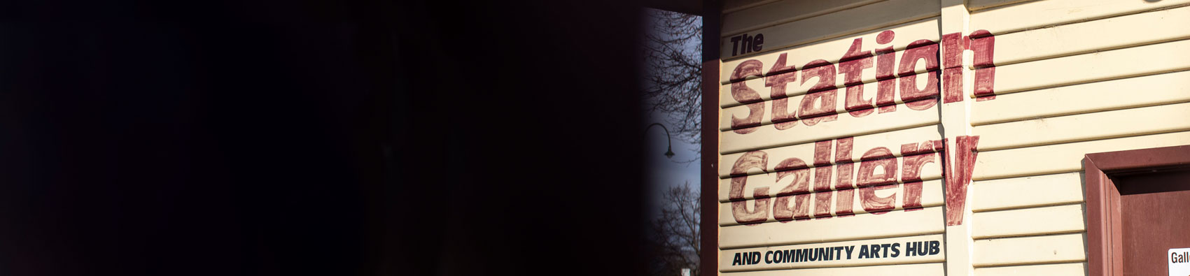 The outside of a wooden building. The layered wooden slabs are painted yellow and there is a sign on the wood saying "Station Galler and community arts hub".