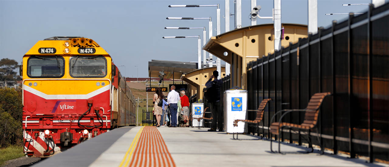 A red and yellow V/Line train sits at the platform of Epsom Station. There are passengers standing on the platform and there are seats, rubbish bins and a passenger information display.