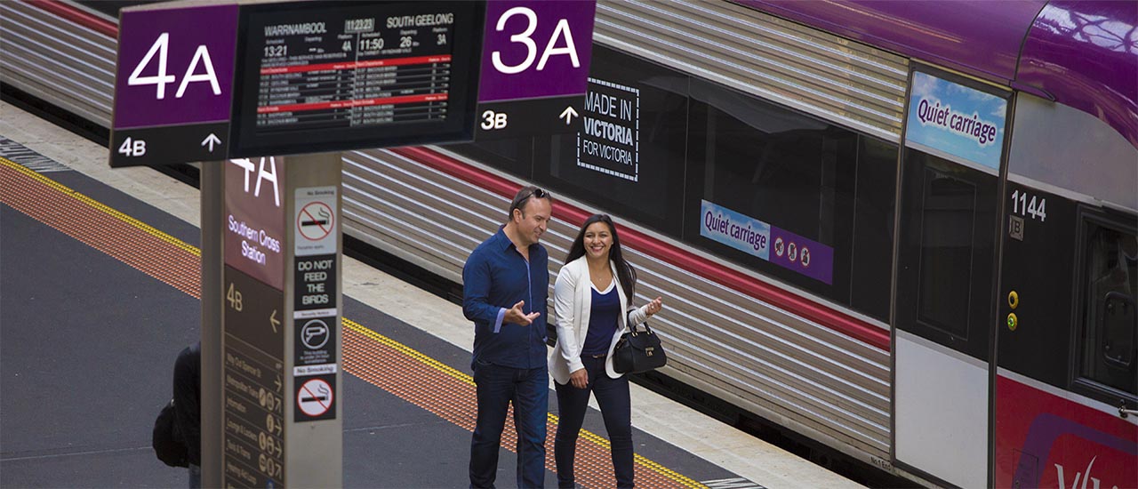 Two passsengers are walking along a V/Line platform at Southern Cross Station. There is a V/Line train in the background, and above the passengers are passenger information displays showing the next scheduled train to depart from the platform.