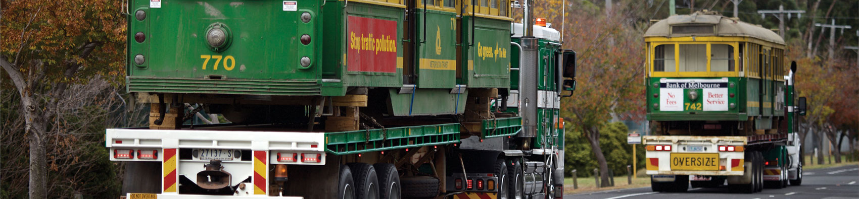 Two heritage W Class trams in yellow and green being transported on the back of two trucks.