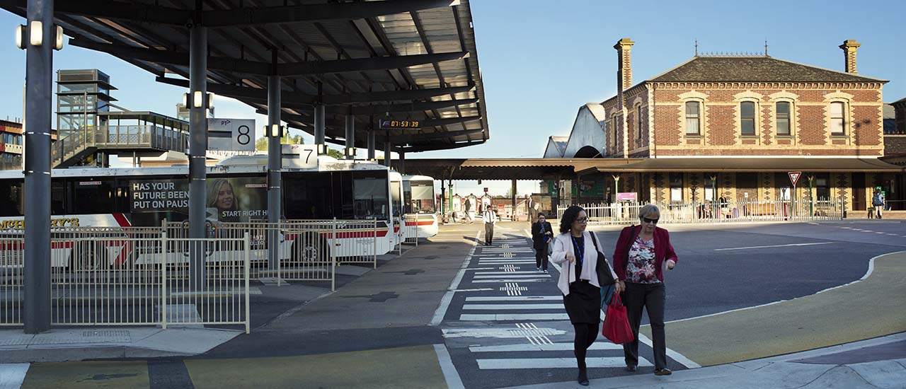 An outdoor view of Seymour Station showing the bus precinct area with several buses parked. Four commuters are crossing the pedestrian crossing which goes past the bus station.