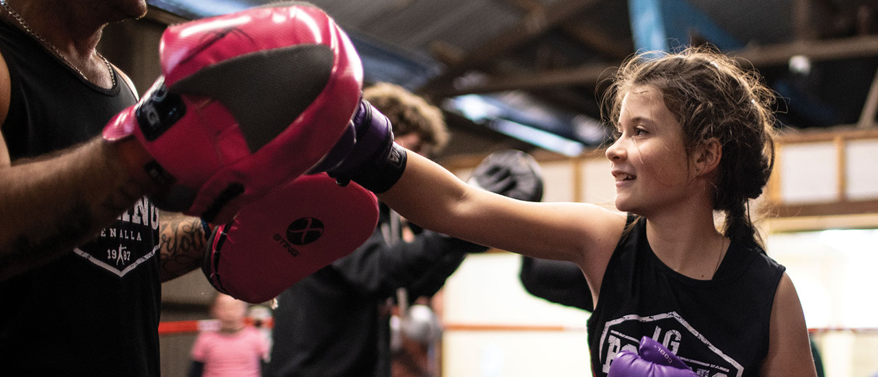 A young child practising boxing in a gym. Child wearing boxing gloves and punching trainer's mits. 