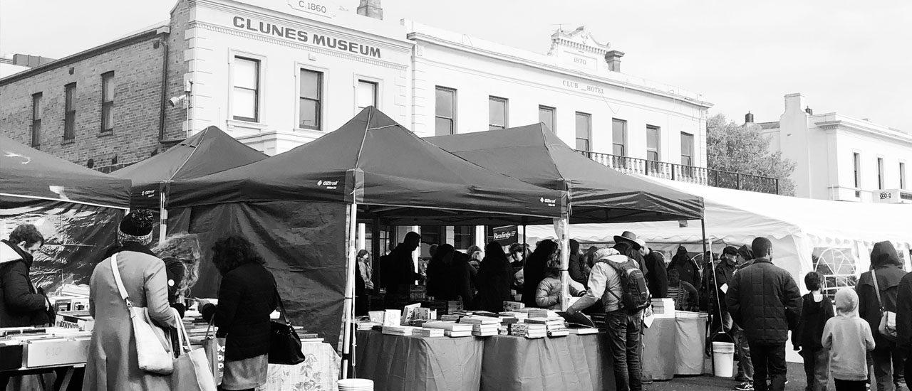 Black and white image of people browsing book stalls in the street during Clunes Booktown festival. Clunes town centre buildings can be seen in the background. 