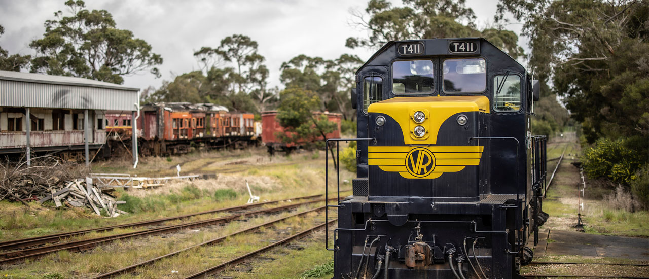 Diesel locomotive T411 painted in Vic Rail blue and yellow livery at Mornington tourist railway. Railway carriages and cloudy sky can be seen in the background. 
