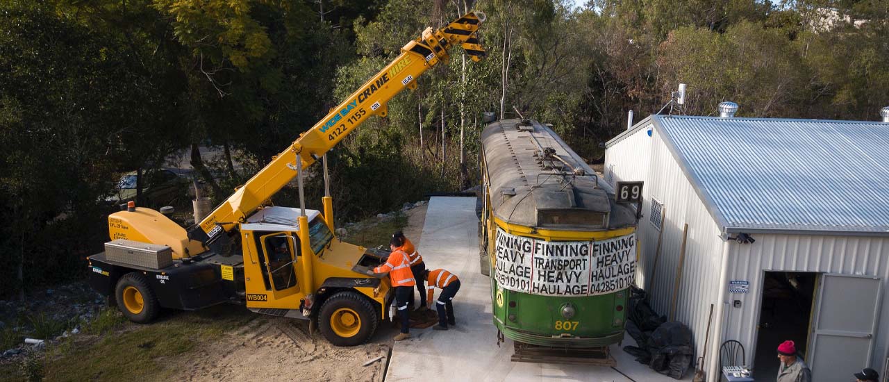 Three mean wearing hi vis safety wear standing next to an orange crane and a green and yellow tram.