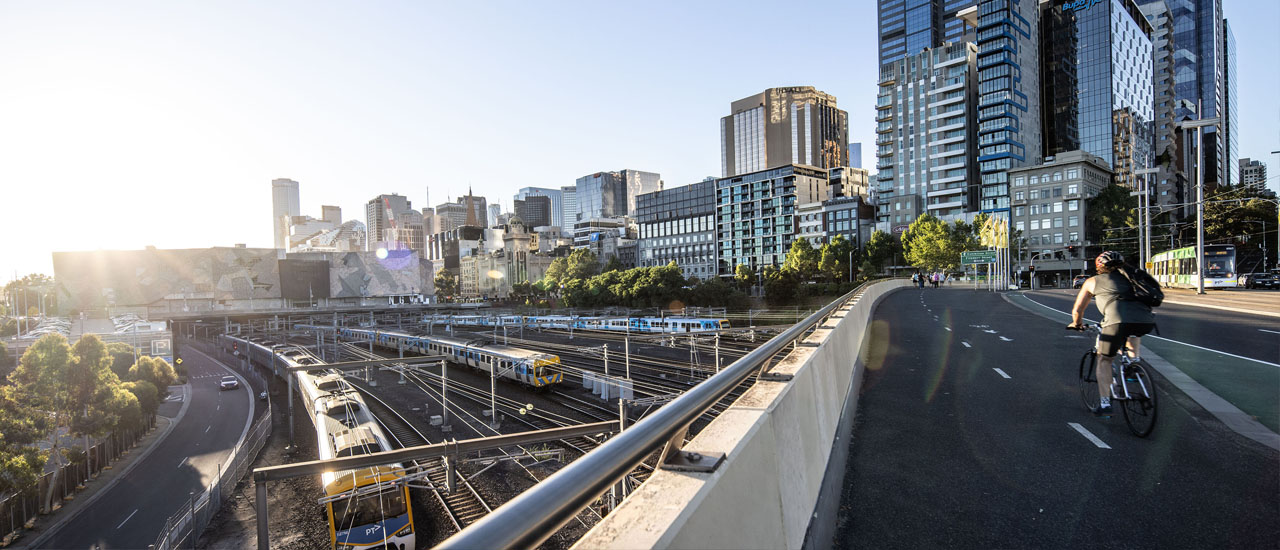 A metro train runs around a bend in front of the Melbourne skyline. A cyclist rides a bicycle across an overpass above the train. A second train, buildings and blue sky can be seen in the background.