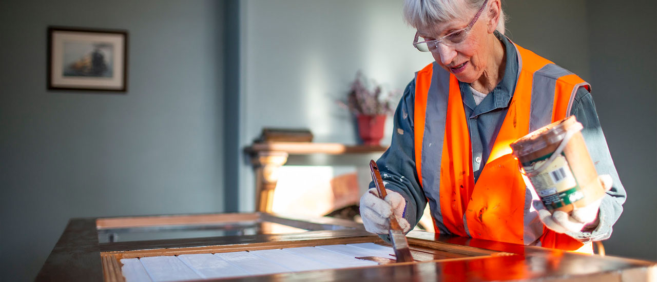 A person wearing a high-visibility vest applies white paint with a brush to a piece of timber. They are working inside a room in  the Newport clocktower building