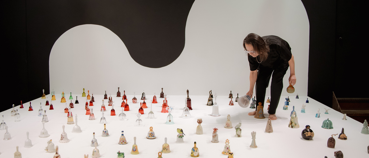 An artist dressed in black moves among  a collection of hand bells of different sizes and colours that are spread out on a white floor. The artist is ringing one bell and holds another in their spare hand. 