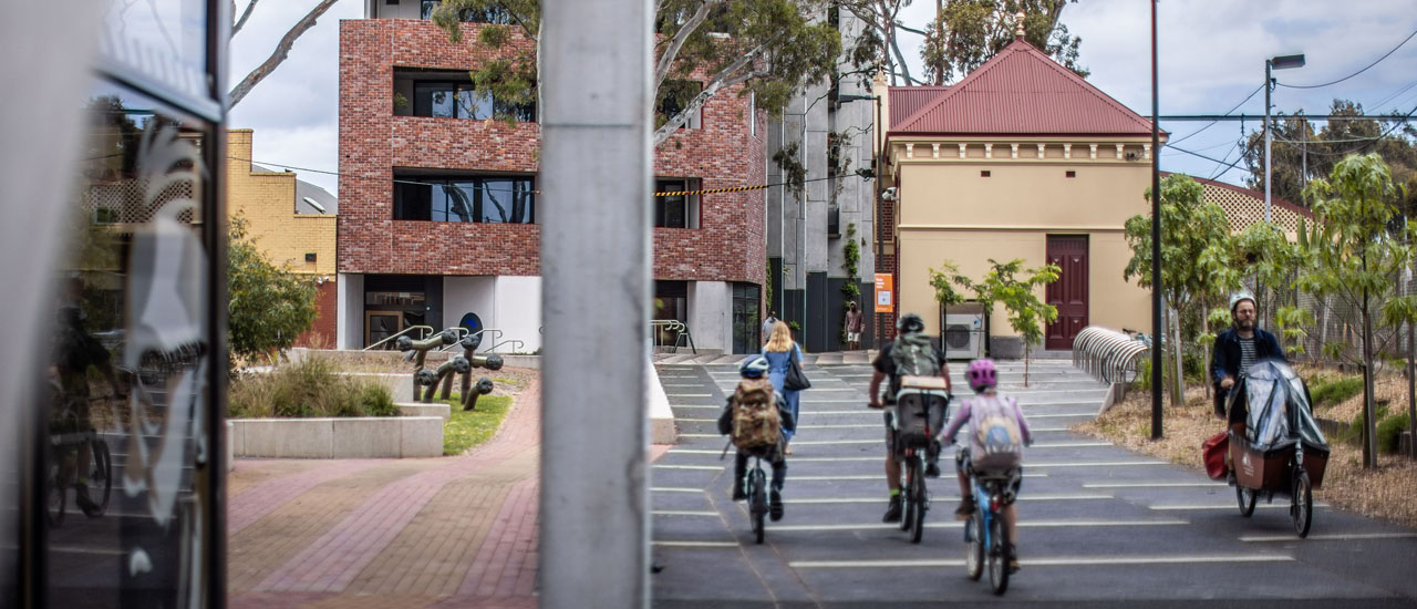 People cycle along a path next to Jewel Station. A red brick apartment development and the old station building can be seen in the distance.