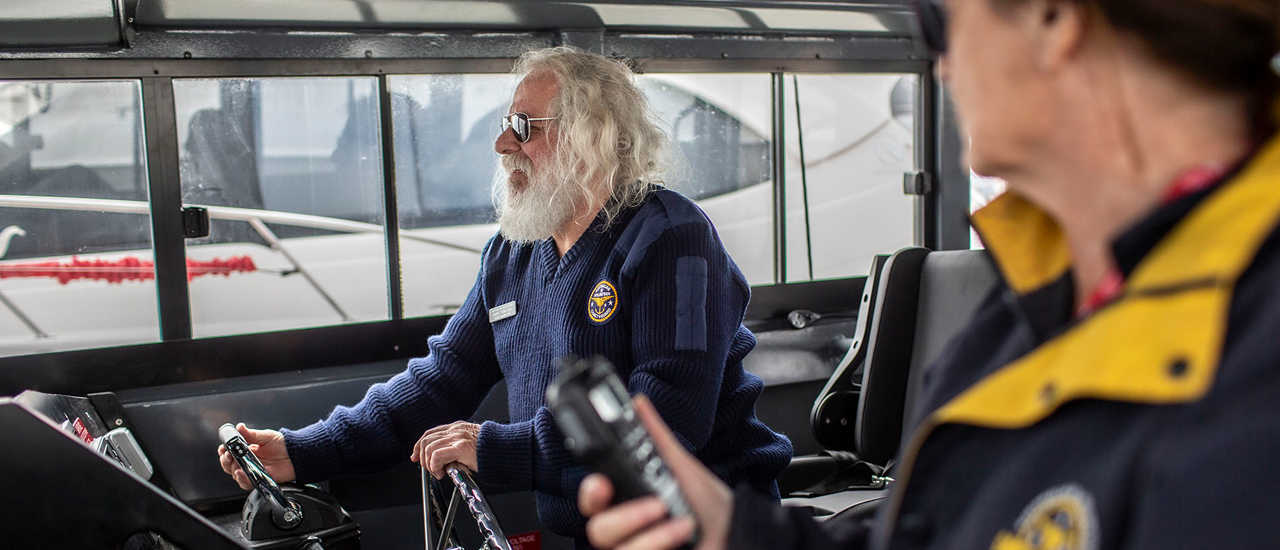 Two people in the wheelhouse of a coast guard boat. One is steering the boat with its wheel while the second person looks on. 