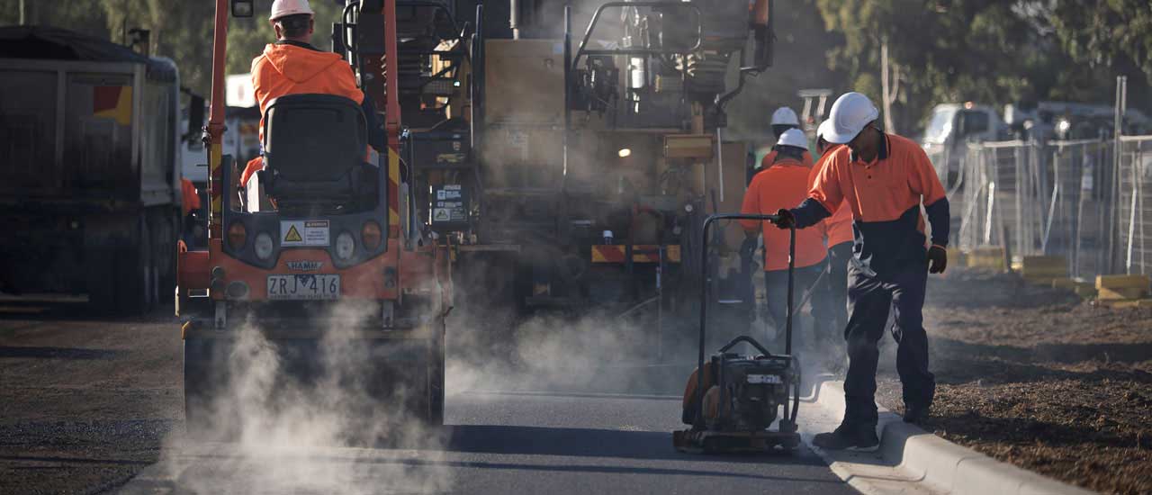 A group of construction workers using machinery to lay asphalt for a new car park. Workers are wearing high vis clothing and hard hats. There is steam around the equipment and workers. .