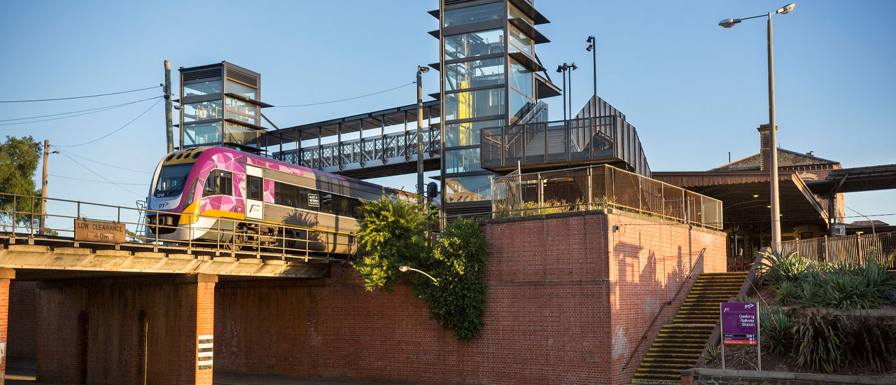 V/Line train crosses a red brick railway bridge. Train station and blue sky in the background. 