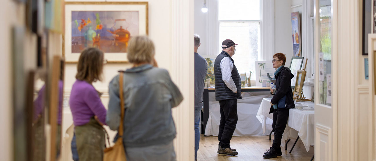 Two people are looking at a painting displayed on the wall inside an art gallery at Woodend Station. Two more people are chatting in the background.