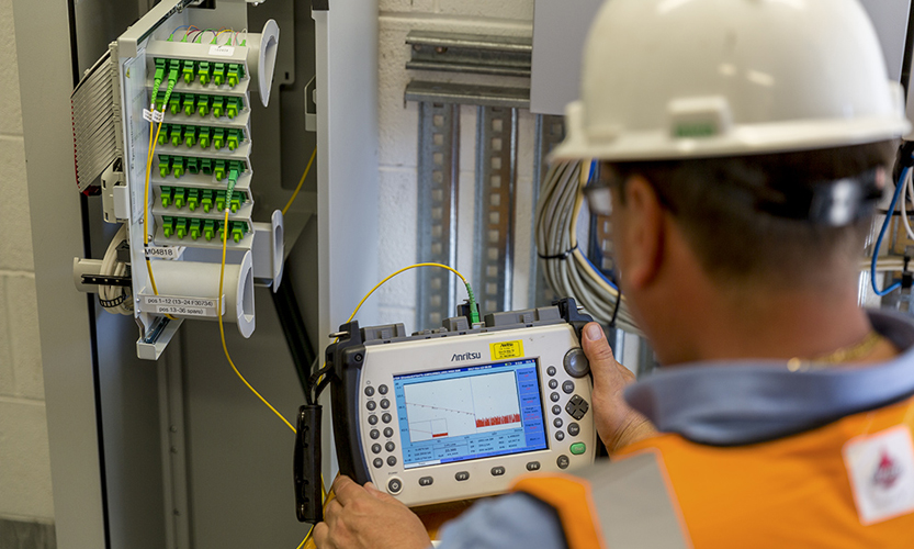 A worker wearing a white hardhat and holding a device to measure telecommunications infrastructure in the background.