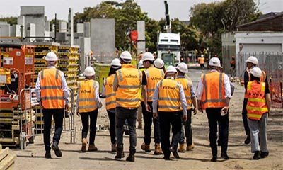 Group of visitors to a construction site walking into the distance wearing high vis and white hard hats.