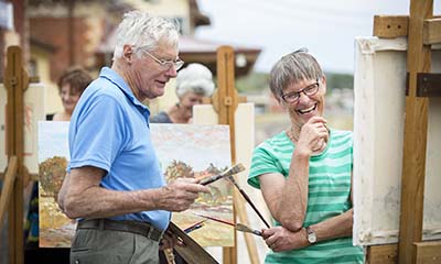 To people admire a painting that is sitting on an easel. They are standing on the platform of Beaufort Railway Station. More people painting can be seen in the background. 