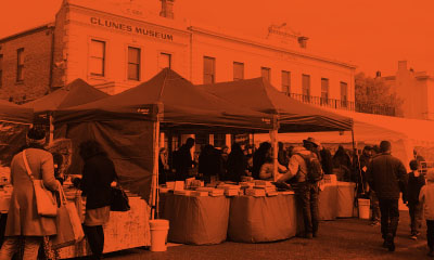 Black and white image  with orange overlay. People browsing book stalls in the street during Clunes Booktown festival. Clunes town centre buildings can be seen in the background.