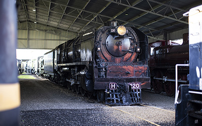 A black heritage steam locomotive on display at an outdoor railway museum. The engine is known as Heavy Harry. It is protected from weather by a roof. 