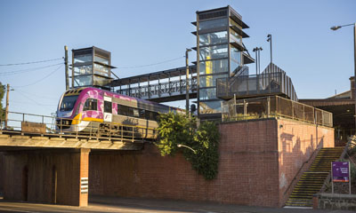 V/Line train crosses a red brick railway bridge. Train station and blue sky in the background.